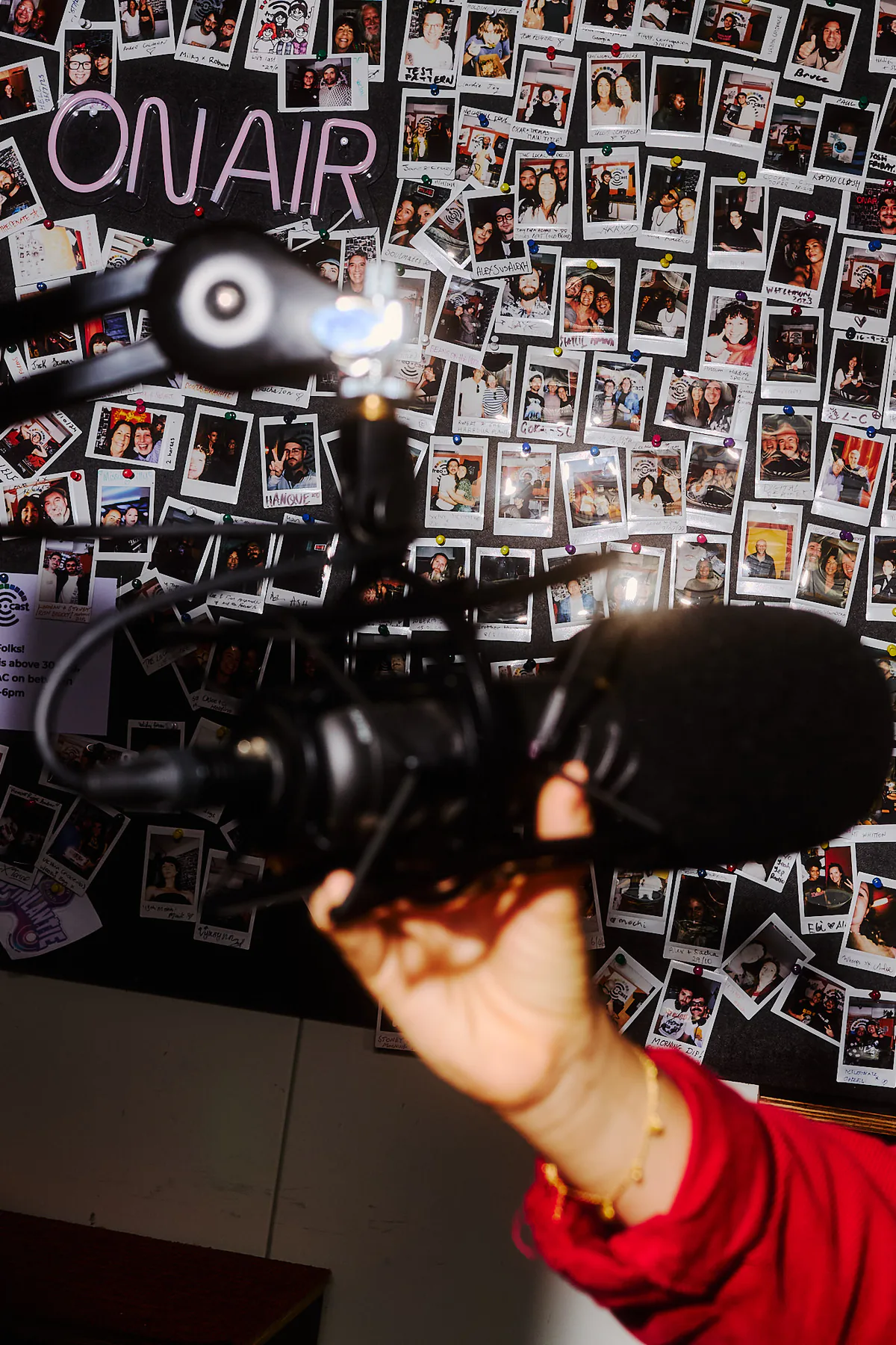 Close-up of a studio microphone and a hand adjusting it, with an On Air sign and community photographs behind